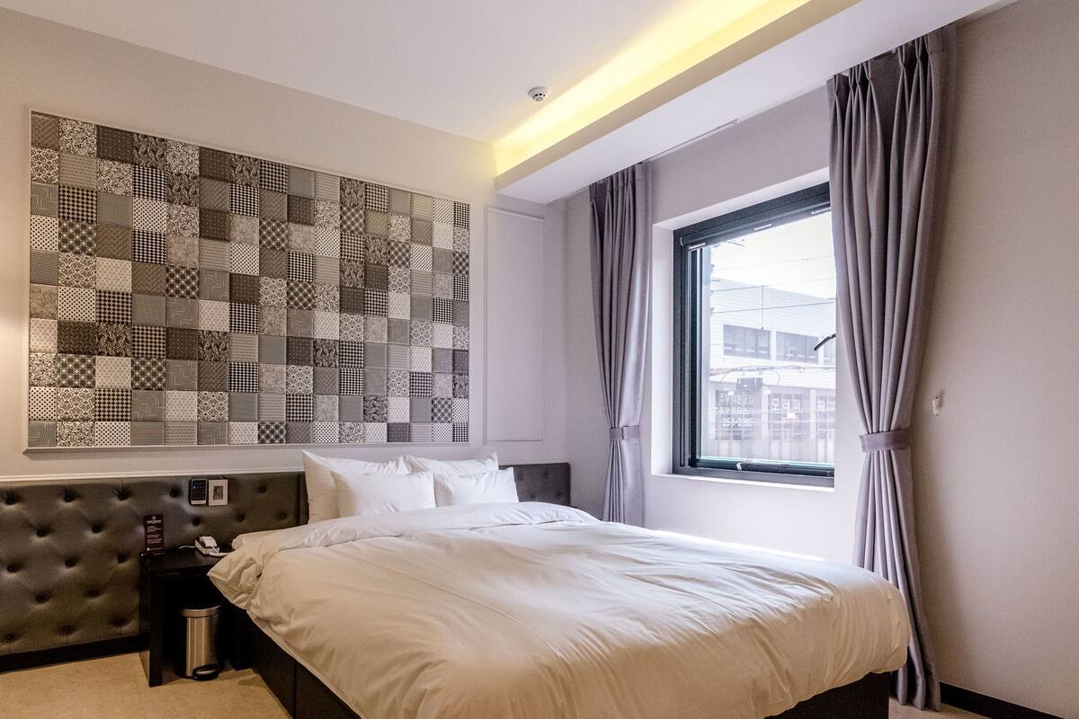Une chambre d’hôtel moderne avec un grand lit, un panneau décoratif au mur, une fenêtre avec des rideaux et une petite table de chevet.
