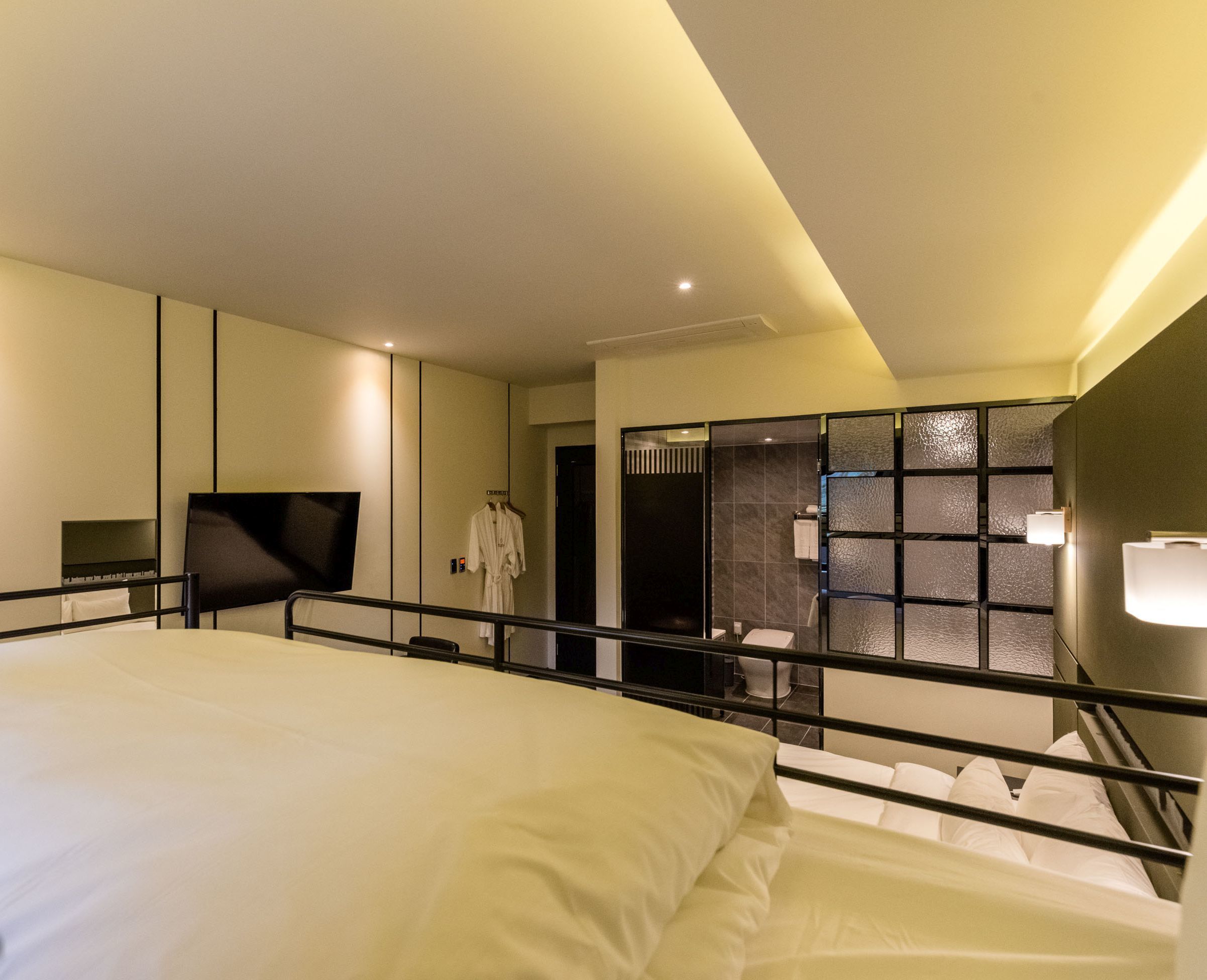 A modern hotel room with a large bed, a flat-screen TV, a robe hanging on a rack, and a bathroom with a glass partition.
