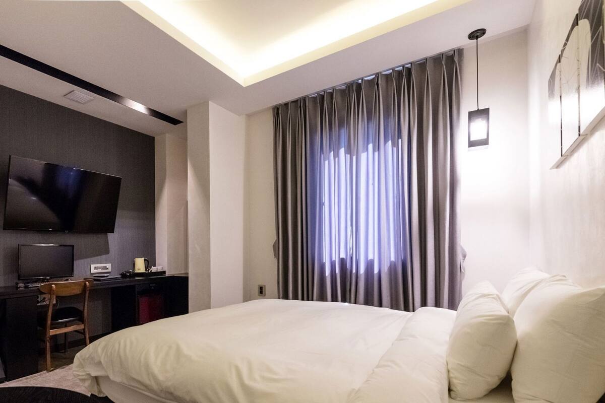 Une chambre d’hôtel moderne dotée d’un grand lit, d’un bureau avec un téléviseur à écran plat et d’une grande fenêtre avec des rideaux.