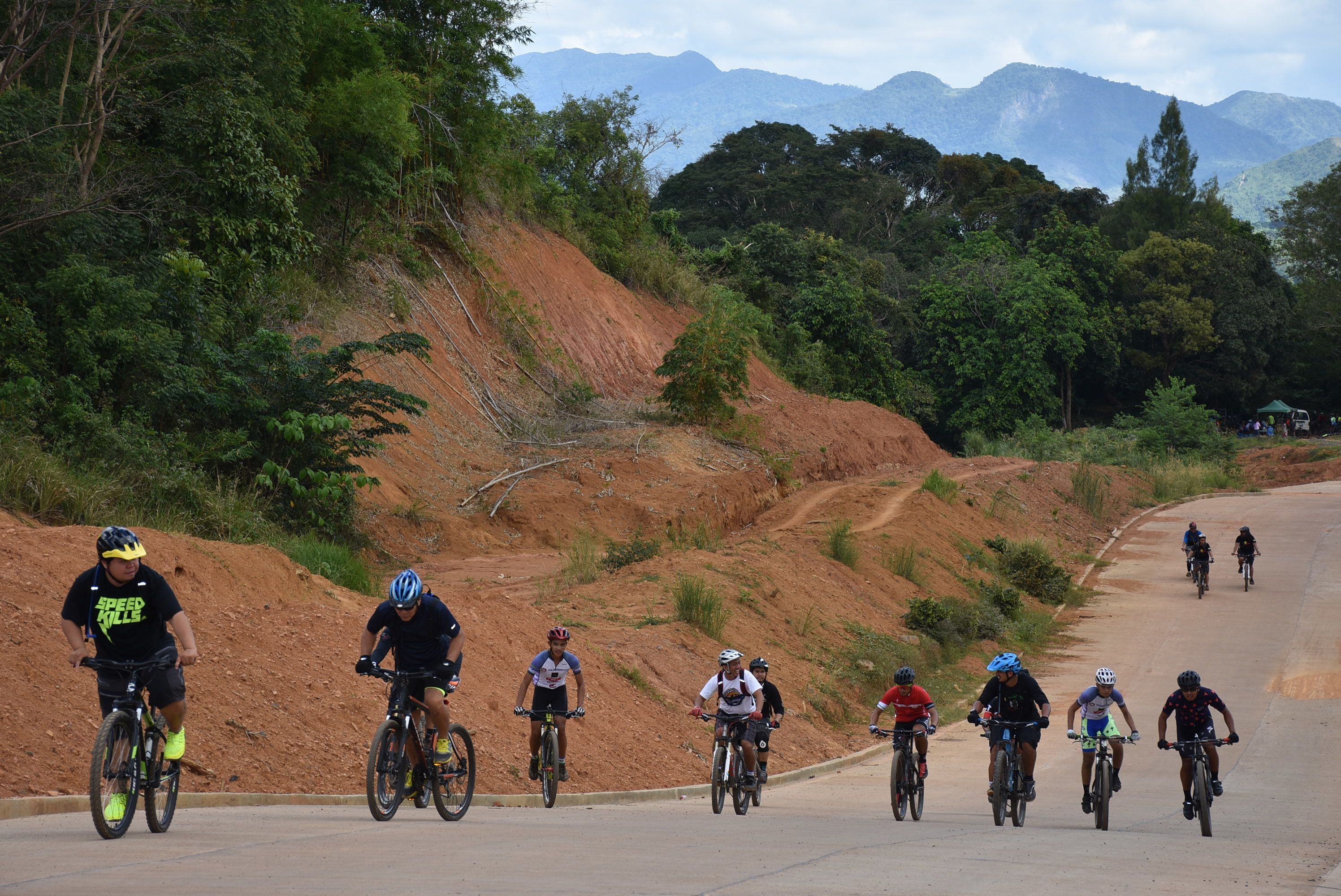 A group of cyclists on a paved road with a dirt path on the side, surrounded by greenery and mountains in the background.