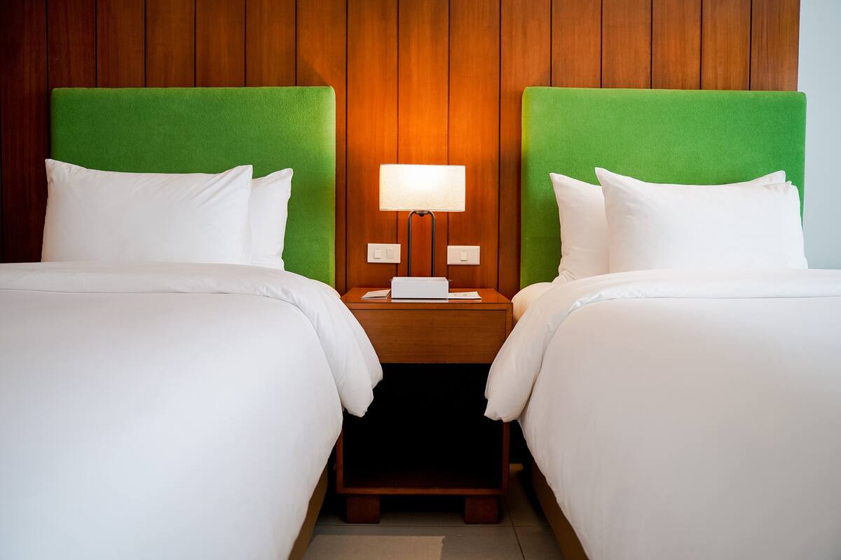 A hotel room with two beds, each with a green headboard, a bedside table with a lamp, and white bedding.