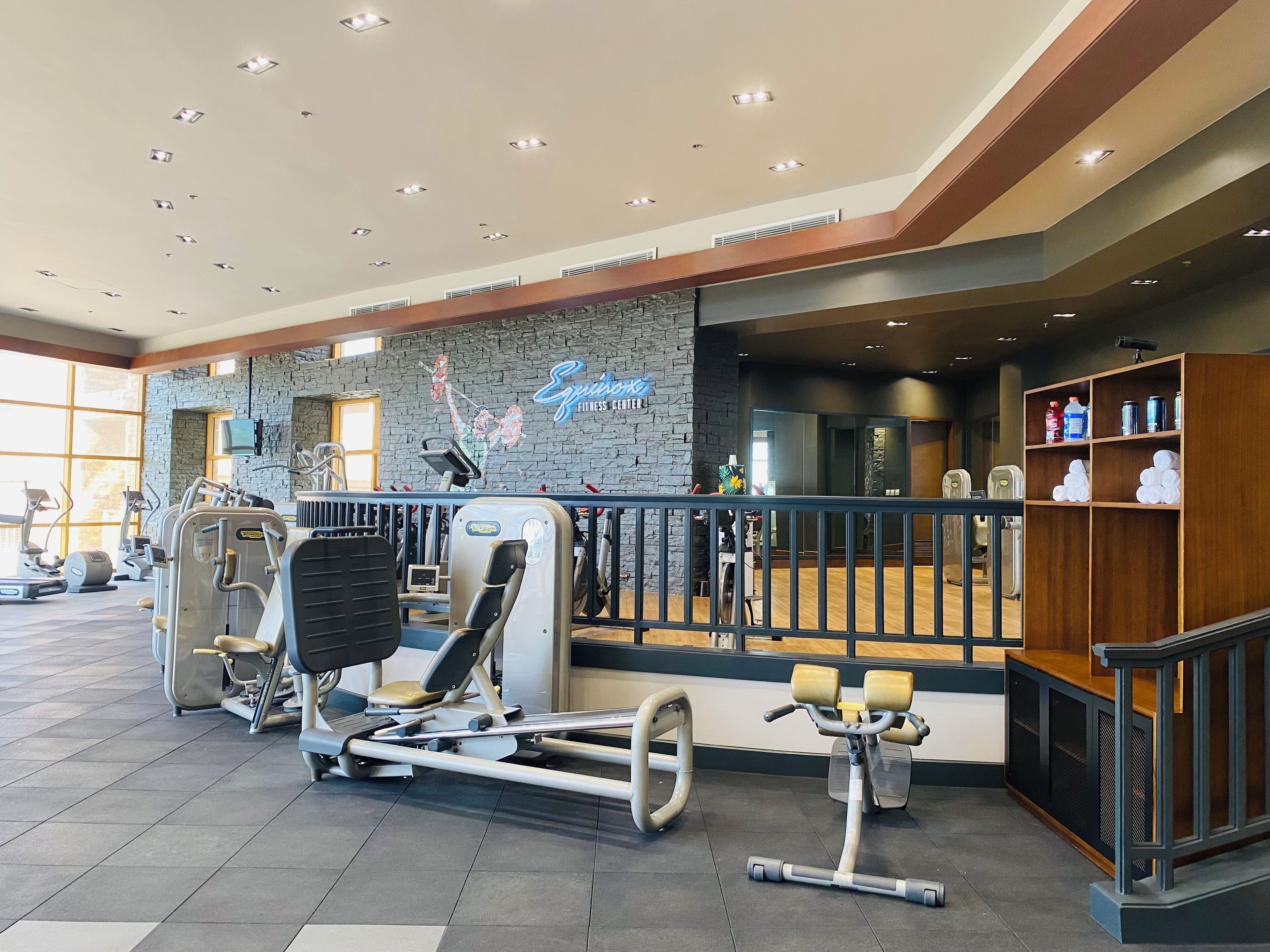 A fitness center with various exercise equipment, a stone wall with a neon sign, and a wooden reception desk.