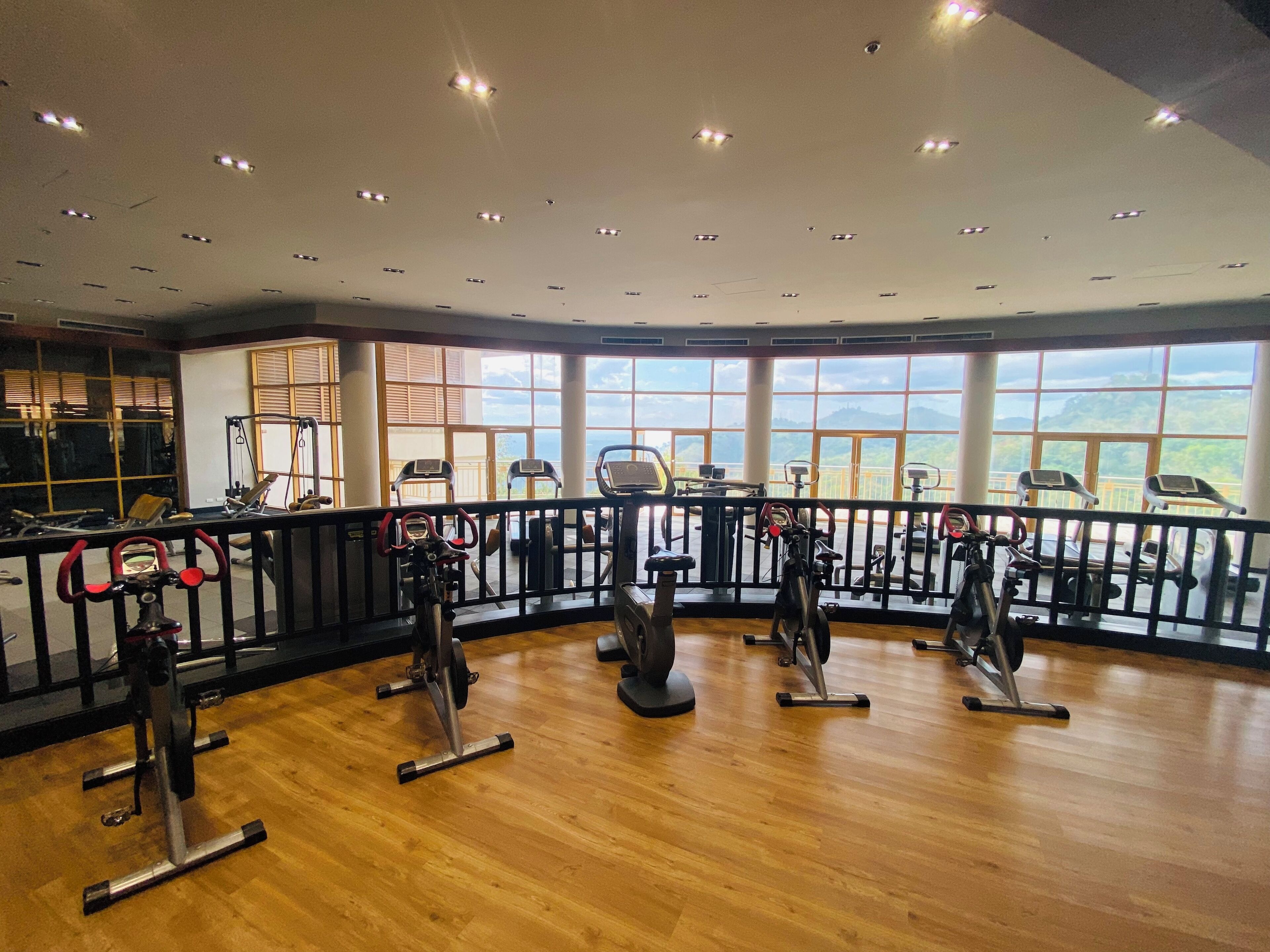Un gimnasio con equipo cardiovascular, ventanas amplias y suelo de madera.
