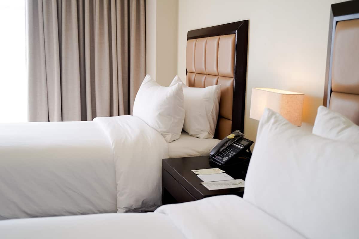 A hotel room with two beds, a nightstand with a phone and papers, and a lamp.