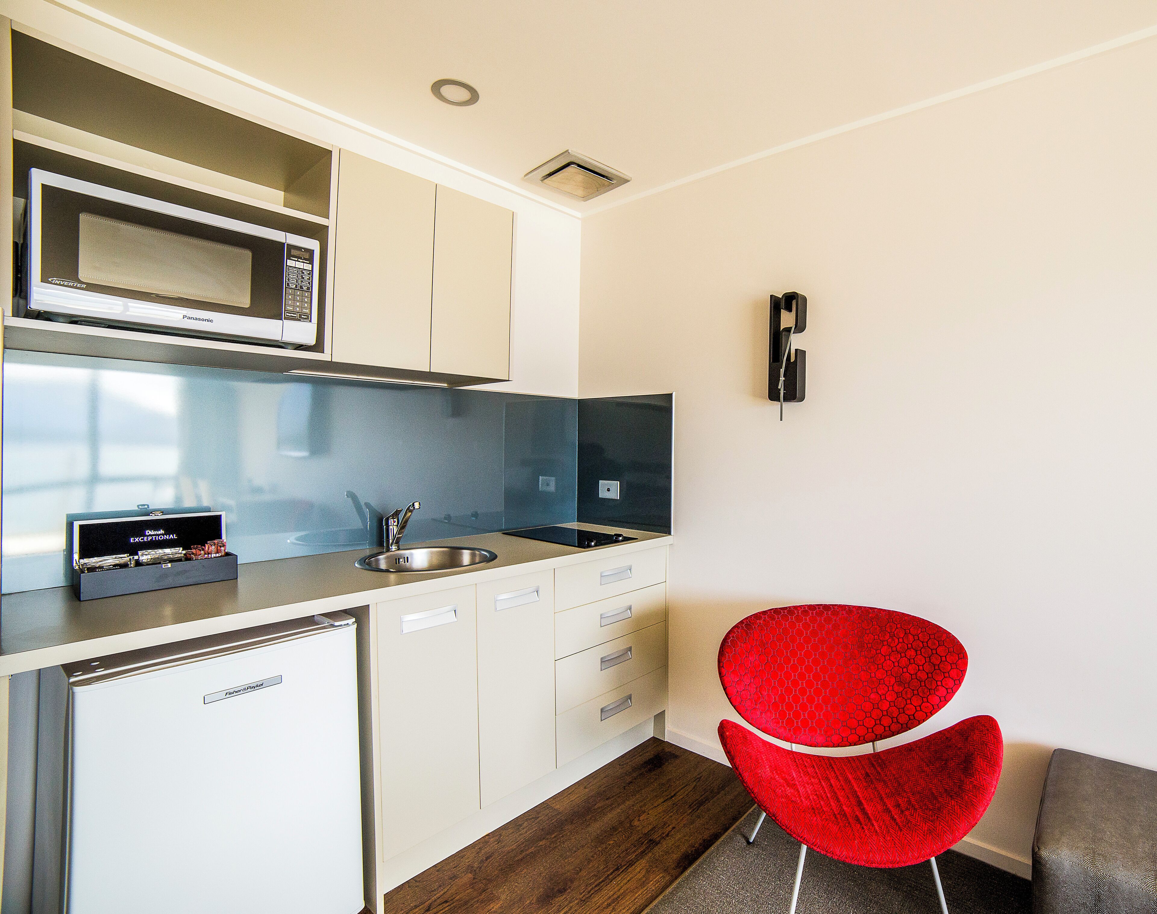 A compact kitchen with a microwave, sink, and a compact refrigerator. There is a red chair and a small table.