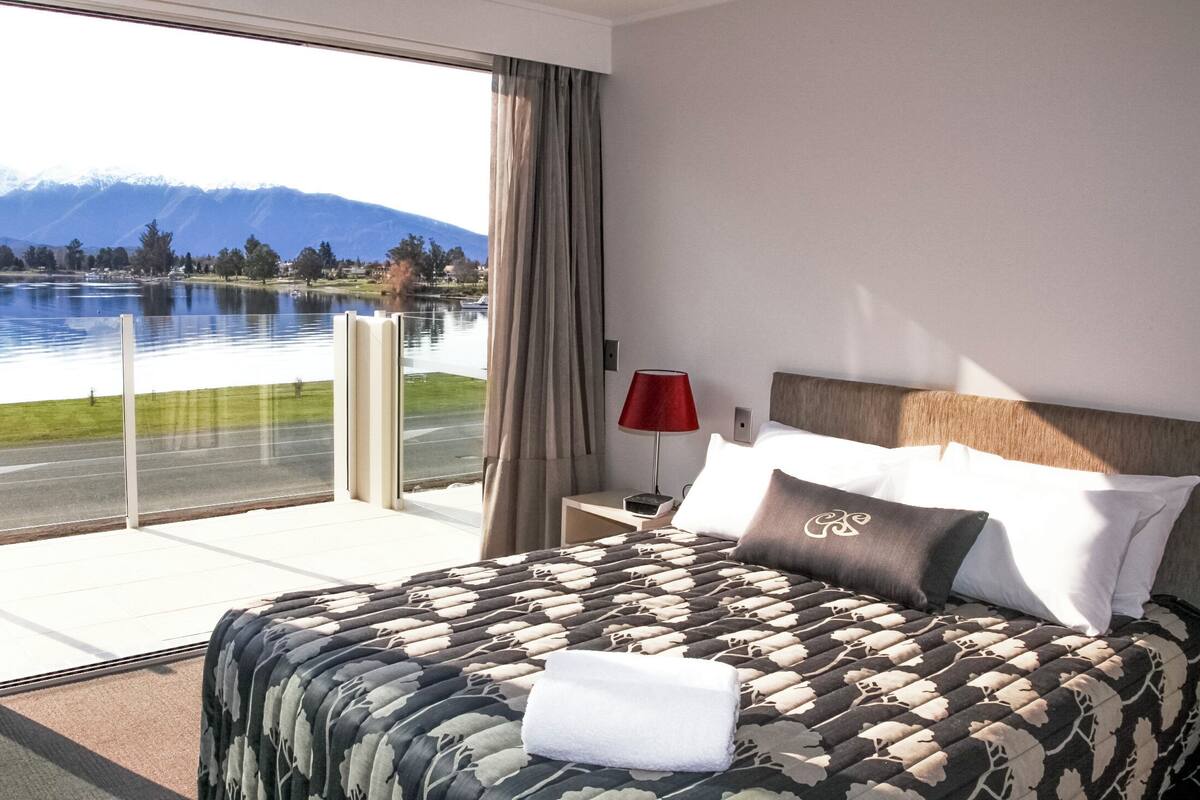 A hotel room with a bed, a nightstand, a lamp, and a view of a lake and mountains.