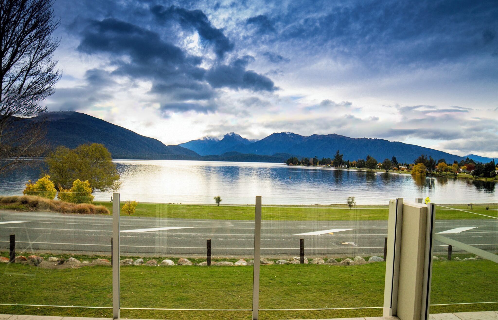 A view of a lake with mountains in the background, a grassy area in the foreground, and a road with a pedestrian crossing.