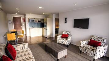 A modern living room with a sofa, ottoman, and floral armchairs, a flat-screen TV, and a kitchen area in the background.