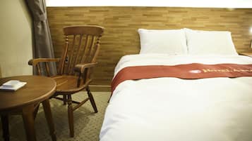 Quarto de hotel com cabeceira de madeira, cama com roupa de cama branca e manta vermelha, uma cadeira de madeira e uma mesa pequena com um objeto branco em cima.