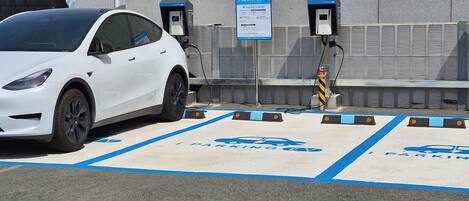 Un coche blanco estacionado en una estación de carga para vehículos eléctricos, con una señal de aparcamiento azul y una caja de carga.