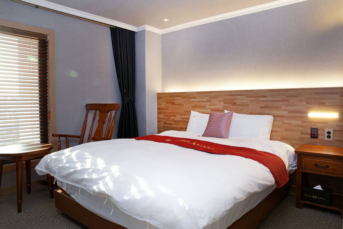 A neatly made bed with white linens and a red blanket, a wooden nightstand with a drawer, a wooden chair, and a window with blinds.