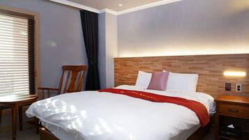 Uma cama bem arrumada com lençóis brancos e um cobertor vermelho, uma mesa de cabeceira de madeira com gaveta, uma cadeira de madeira e uma janela com persianas.