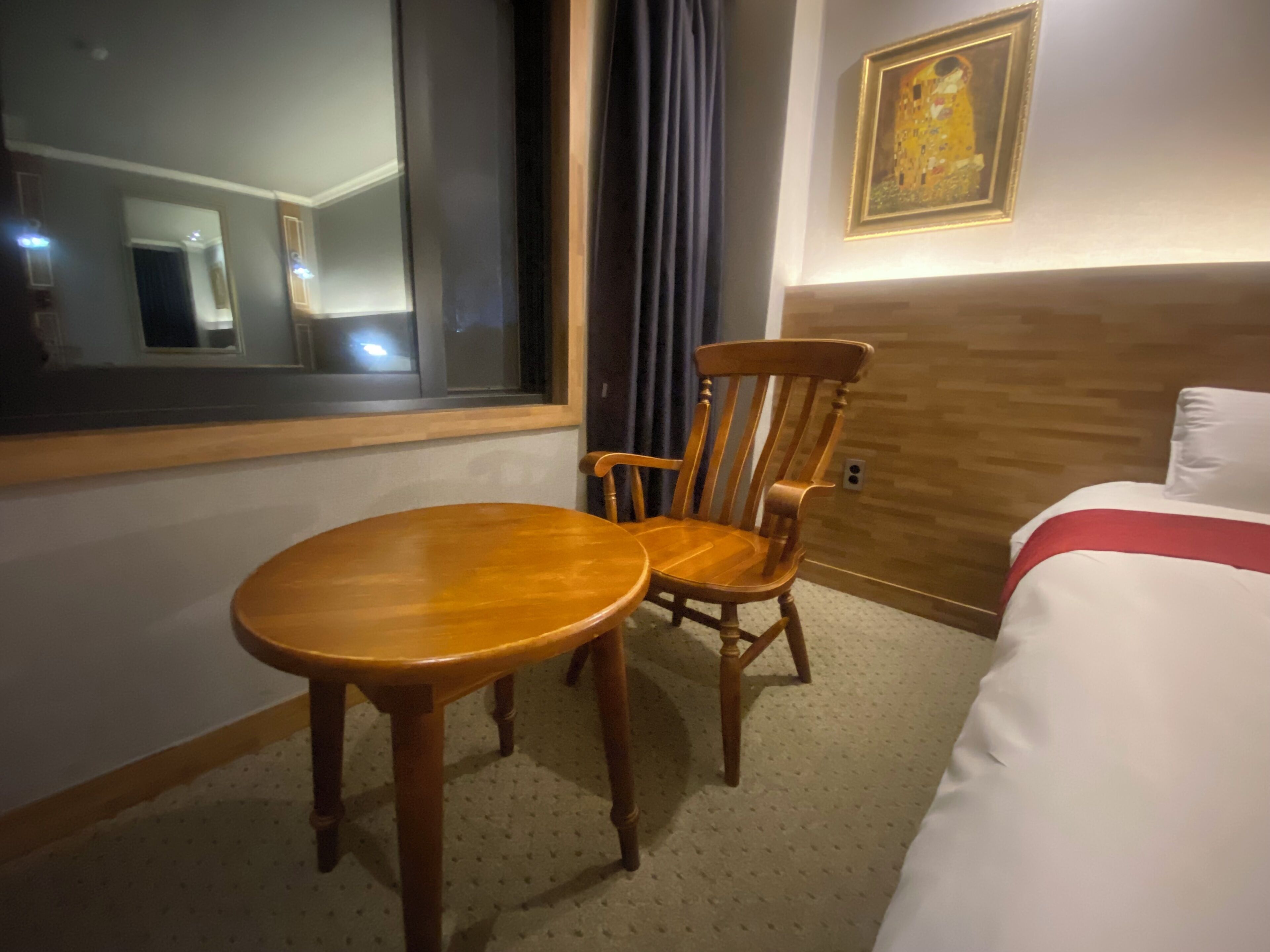 A hotel room with a wooden chair, a small round table, a bed with white bedding and a red accent, and a painting on the wall.