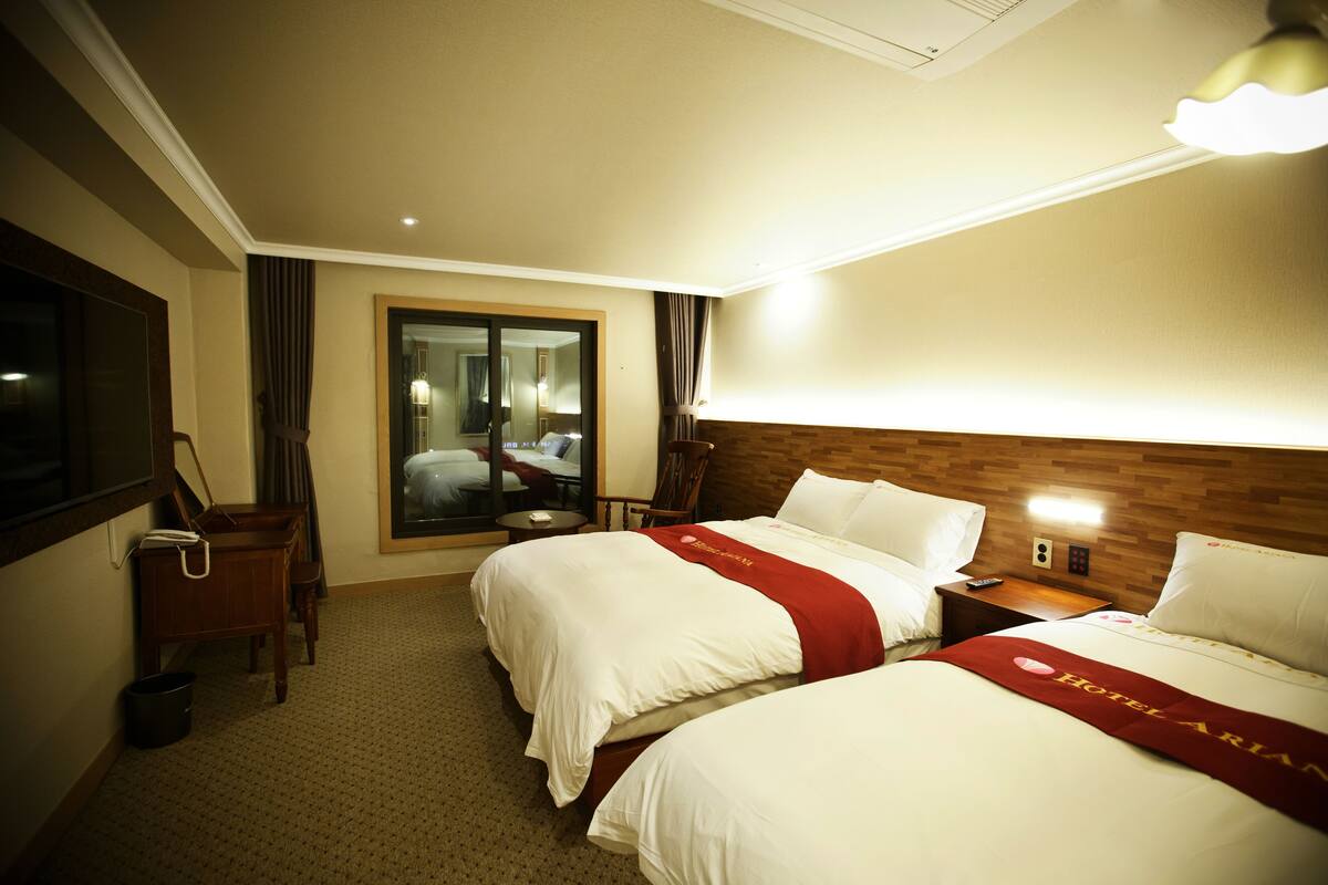 A hotel room with two beds, a desk, a chair, a mirror, and a window.