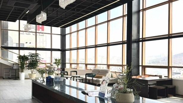 A modern reception area with a marble counter, seating, potted plants, and large windows offering a city view. - Ariana Hotel (Daegu)