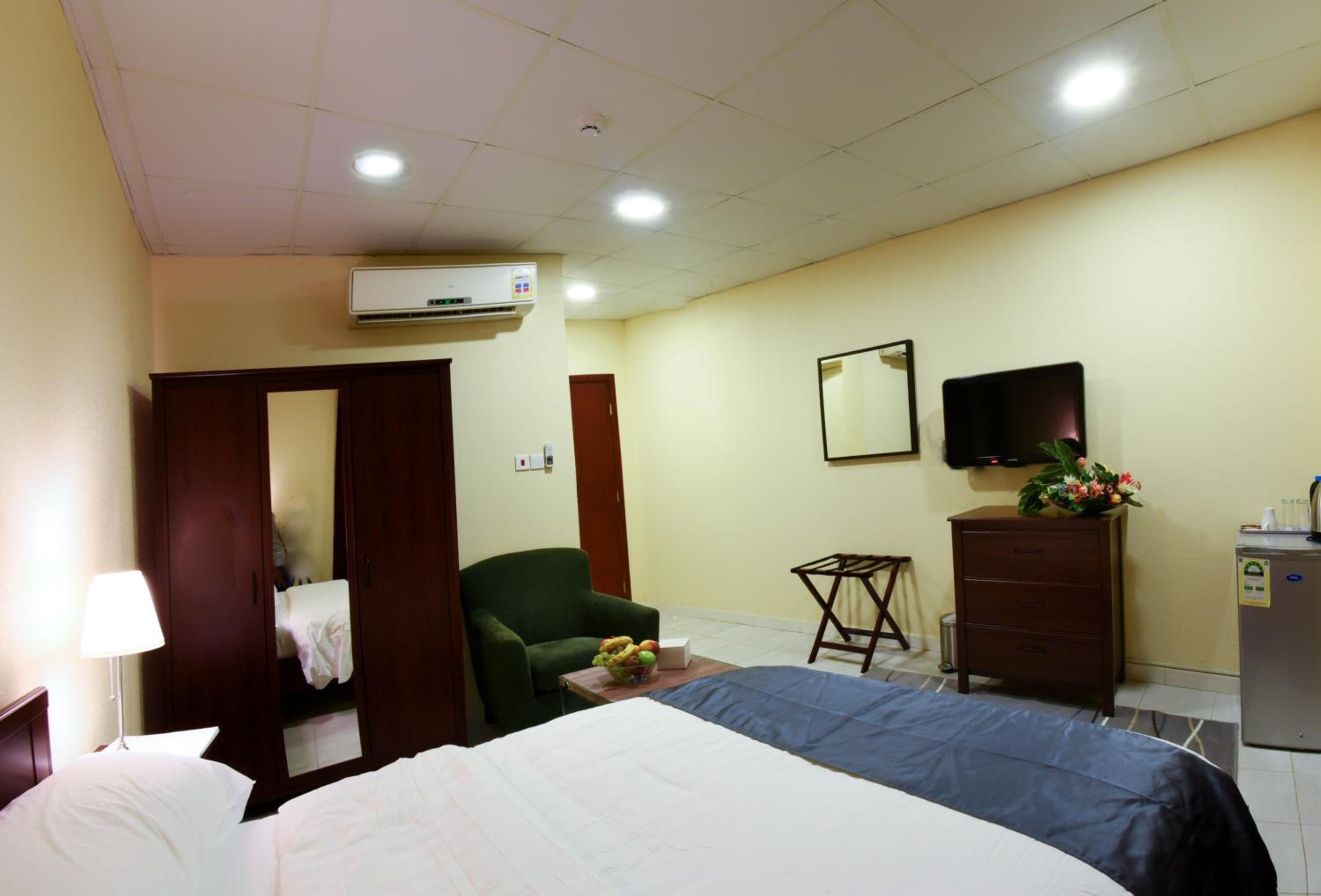 A hotel room with a bed, a chair, a desk, a dresser, a mirror, a television, and an air conditioning unit.