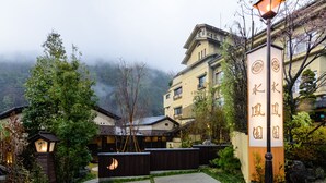 Um edifício japonês tradicional com uma placa, cercado por vegetação e uma montanha enevoada ao fundo.