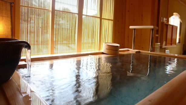 Uma onsen japonesa tradicional com interior de madeira, uma bacia de pedra e um deck de madeira.