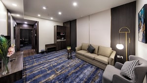 A modern living room with a sofa, armchair, and a coffee table. There is a TV mounted on the wall and a lamp on a side table. - Lily Hotel Suite Hofuf (Al-Hofuf)
