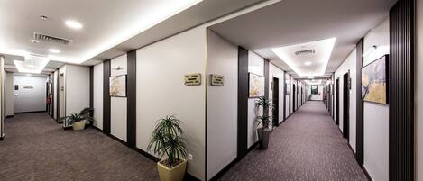 A long, narrow hallway with office cubicles on both sides, potted plants, and signs indicating directions.