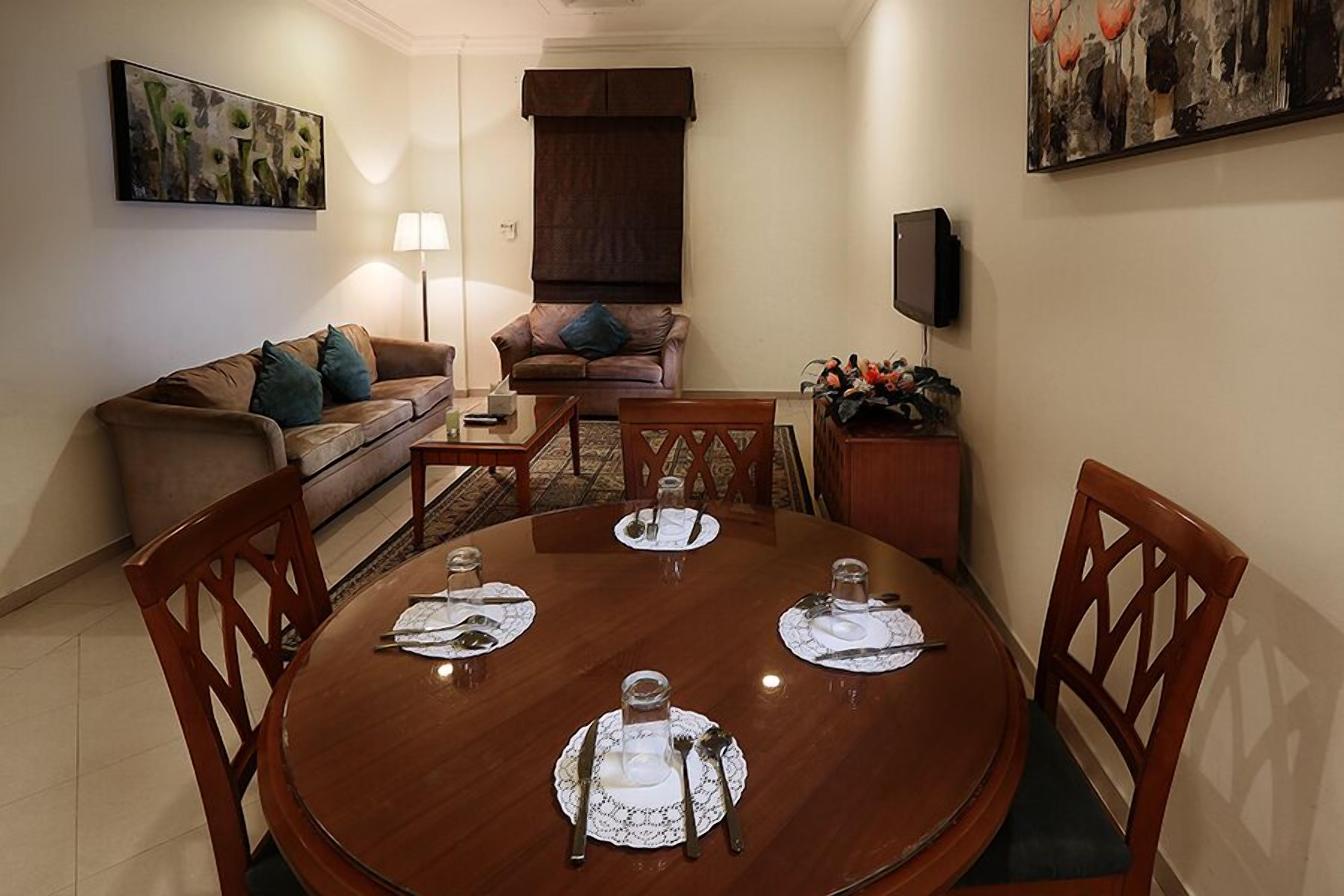 A dining area with a round table set for four, surrounded by wooden chairs. A sofa and a small side table with decorative items are in the background.