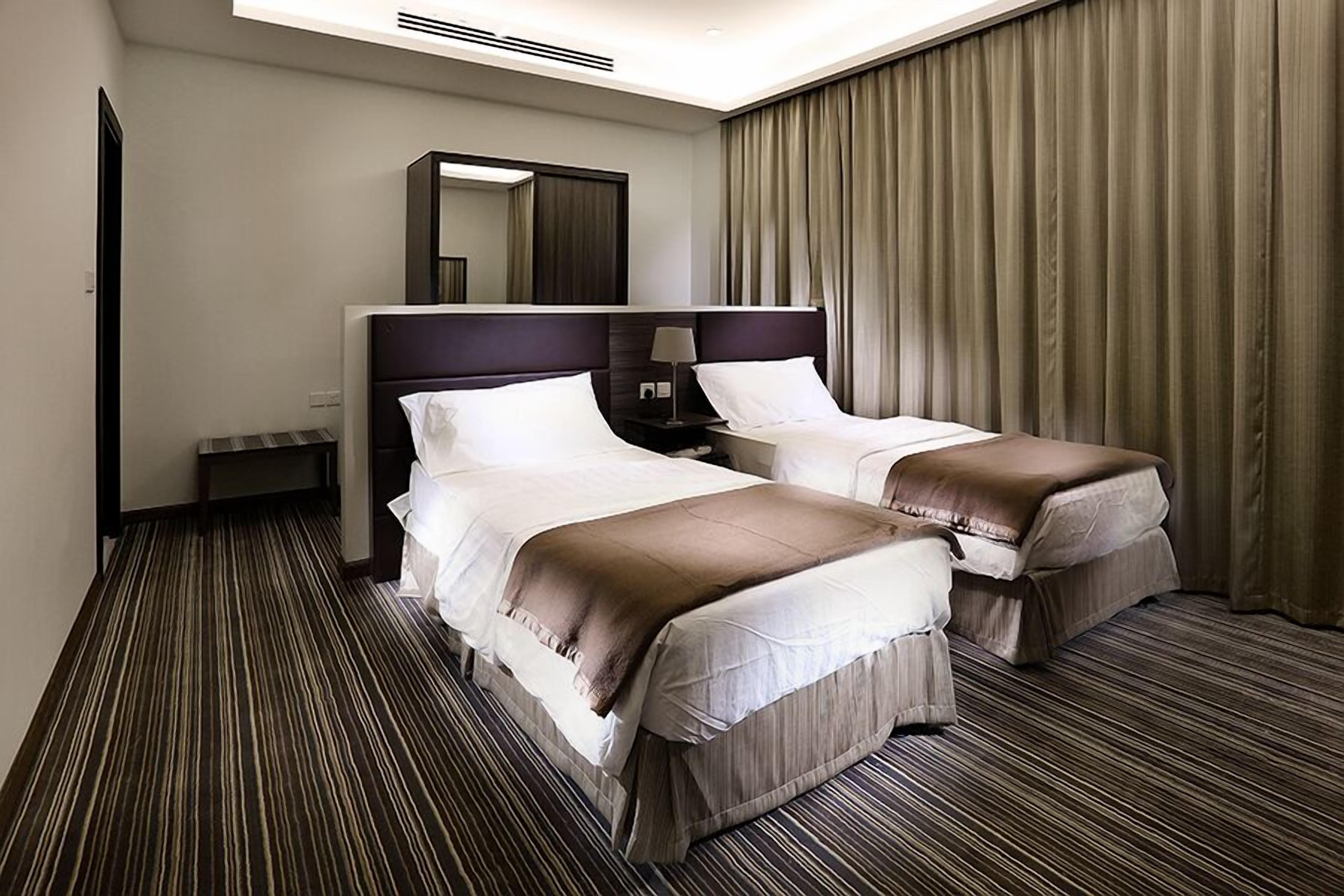 Habitación de hotel con dos camas, un espejo grande, una mesita de noche, una lámpara y una ventana con cortinas.
