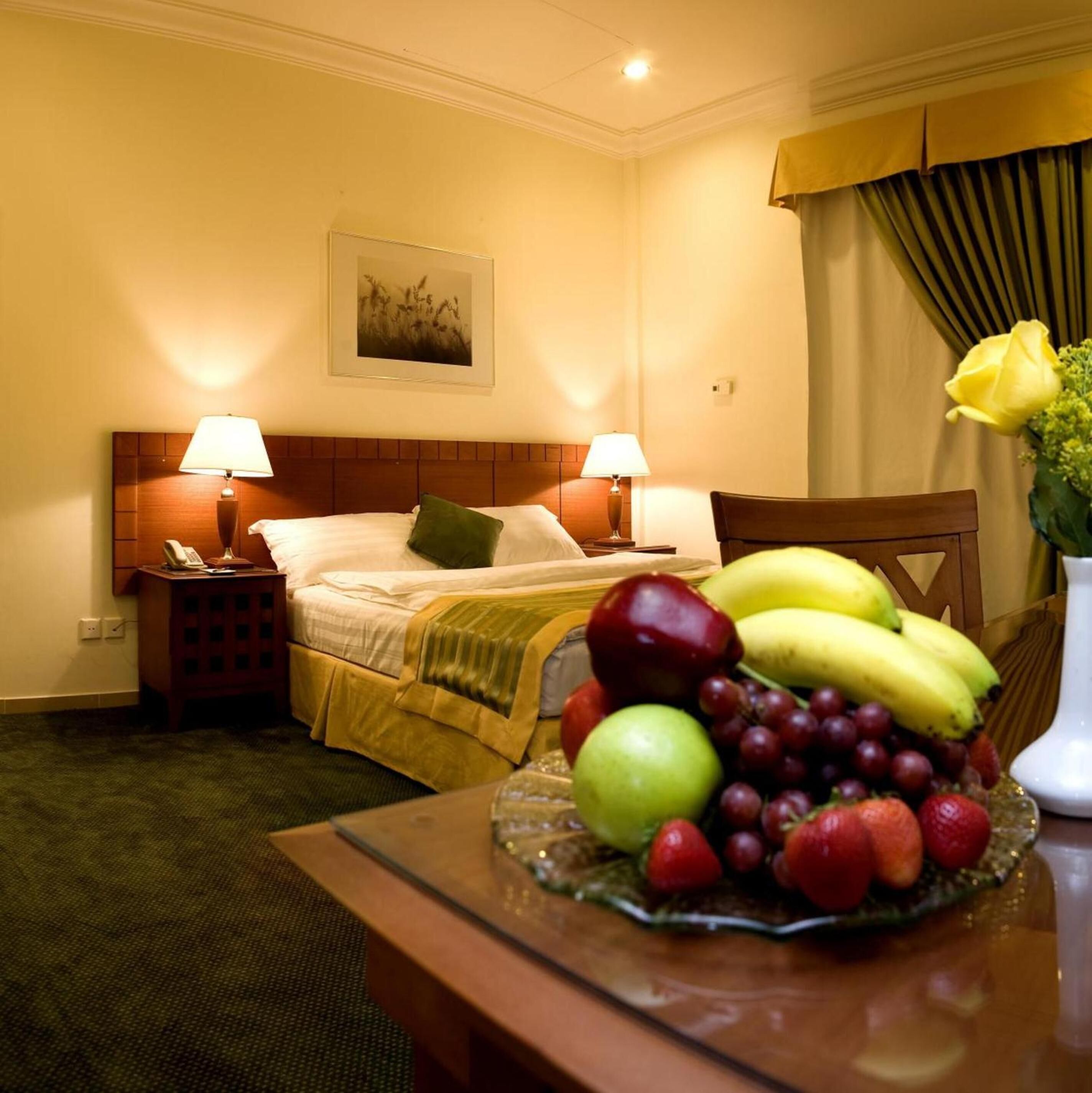 A hotel room with a bed, two bedside lamps, a nightstand, a telephone, a framed picture on the wall, a table with a fruit platter, and a vase with a yellow flower.
