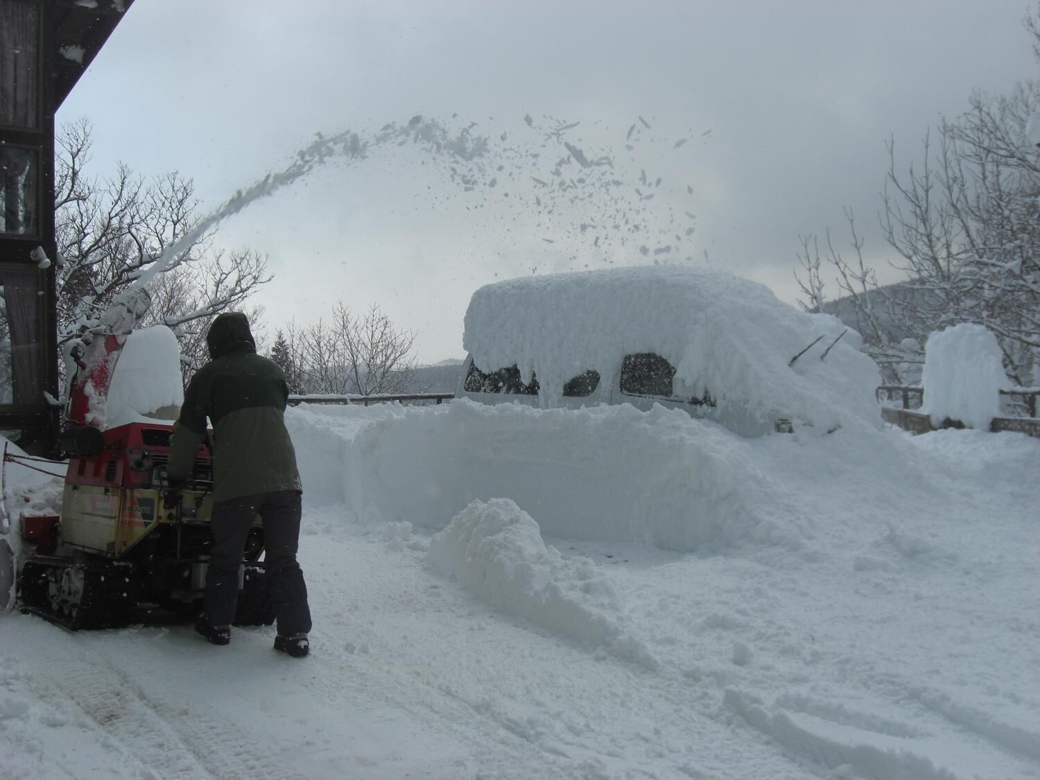 A person operating a snowplow in a snowy environment with large snowbanks and a mountainous backdrop.