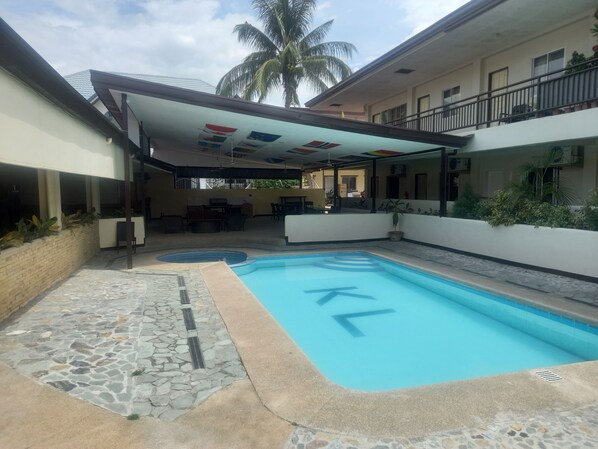 Una piscina con un deck de baldosas, una zona de estar techada y una palmera al fondo.