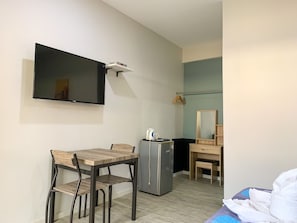Habitación de hotel compacta con cama, escritorio con silla, refrigerador pequeño, televisor de pantalla plana montado en la pared y espejo sobre el escritorio.