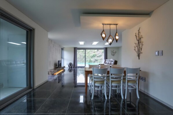 A modern dining area with a large table and chairs, a chandelier, and a view through a glass door.