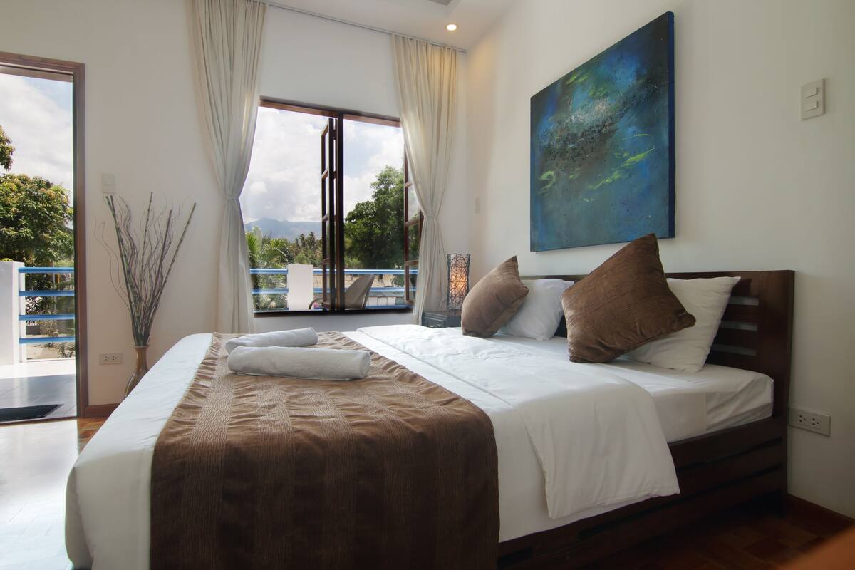 A bedroom with a large bed, a view of the outdoors through a sliding door, and a painting on the wall.