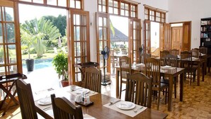 A dining area with wooden tables and chairs, large windows, and a view of a swimming pool and greenery.
