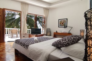 A hotel room with a bed, a TV, a desk, and a balcony with a view of palm trees.
