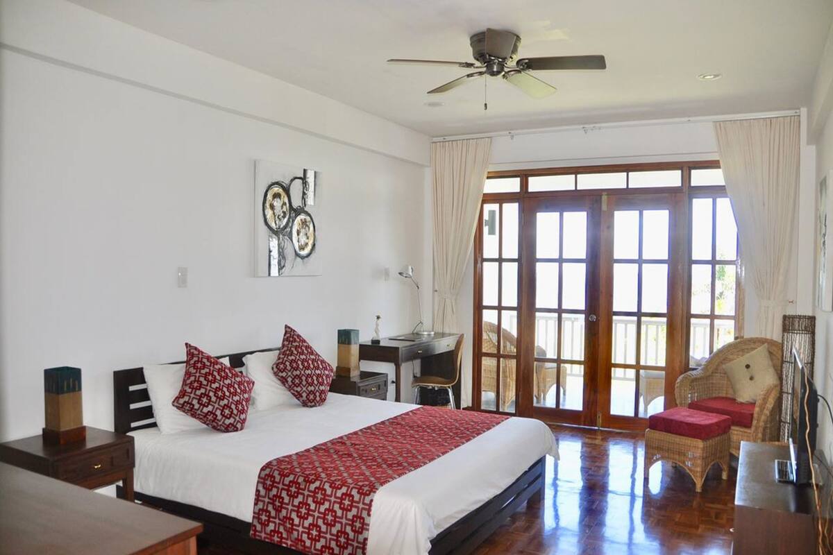 A bedroom with a large bed, a desk, a chair, and a balcony.