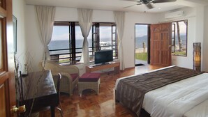 A hotel room with a bed, a desk with a television, a chair, and large windows offering a view of the sea.