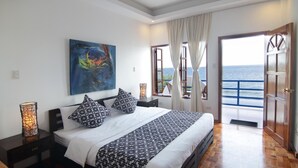 A bedroom with a large bed, a painting on the wall, and a view of the sea through an open door.