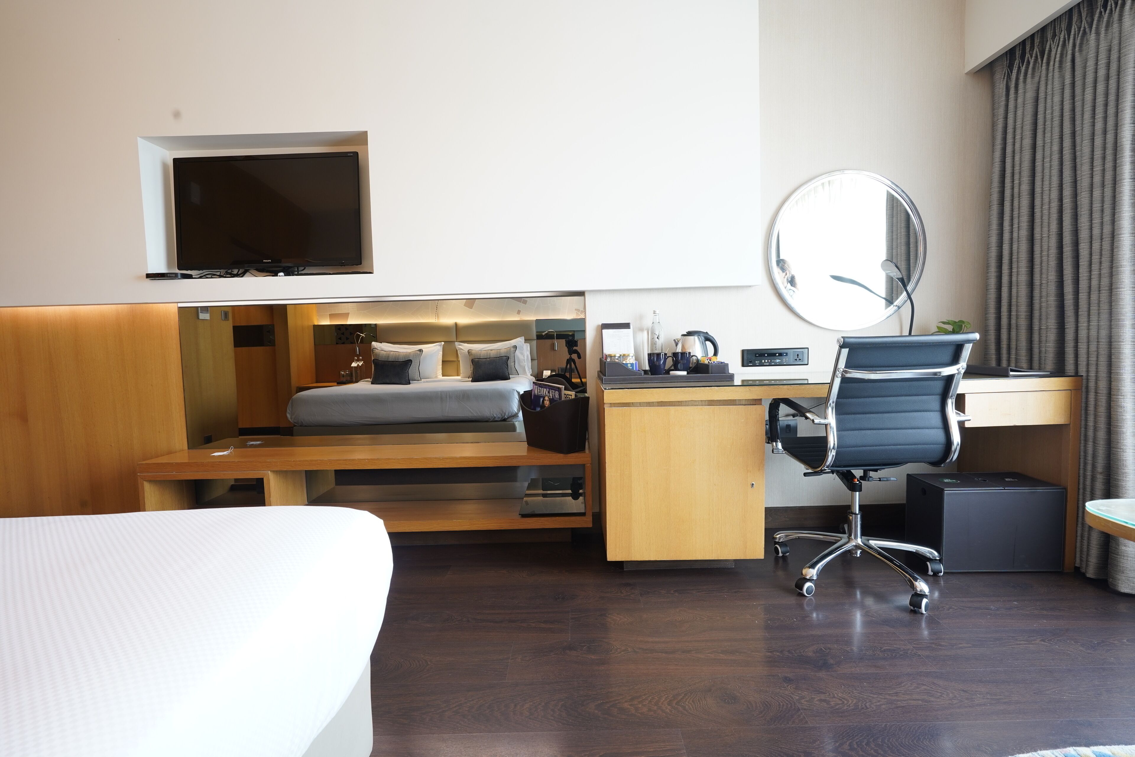 Una habitación de hotel moderna con cama, escritorio, silla, televisor y un espejo grande.