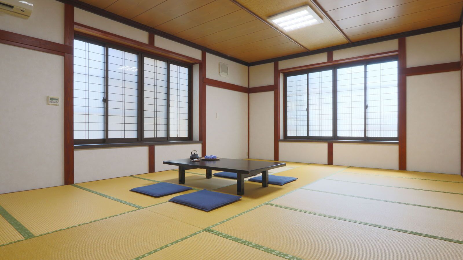A tatami mat room with a table and cushions, large windows, and traditional wooden beams.