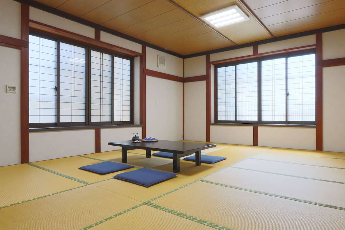 Una habitación con suelo de tatami, una mesa, cojines, ventanas amplias y vigas de madera tradicionales.