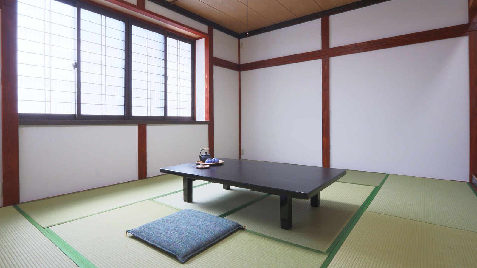 A tatami mat room with a low table and a cushion, a window with grid pattern, and wooden beams.