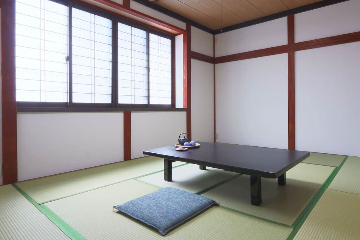 A tatami mat room with a low table and a cushion, a window with grid pattern, and wooden beams.