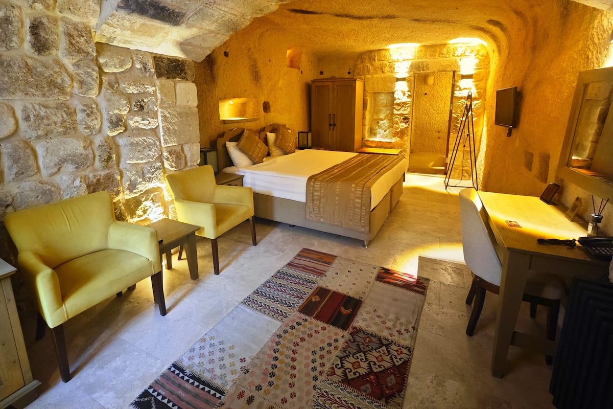Una habitación con paredes de piedra, una cama, sillones amarillos, un escritorio y una alfombra estampada.