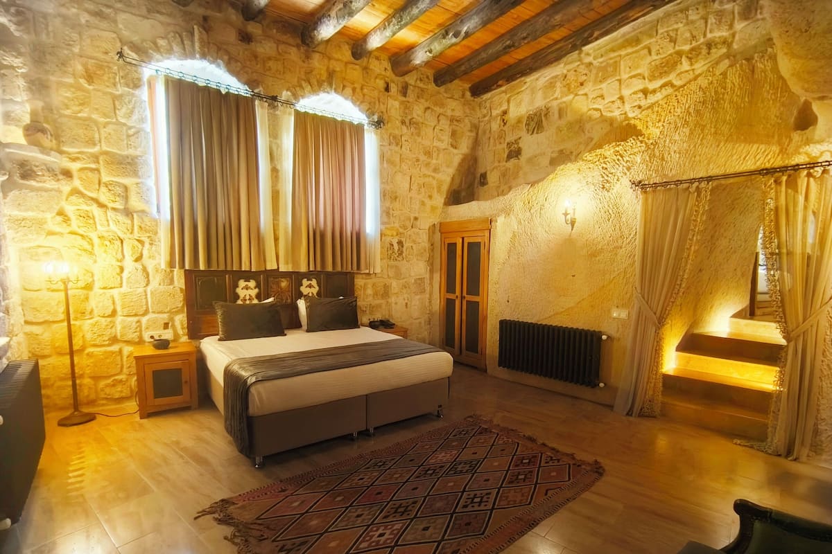 Dormitorio con paredes de piedra, una cama, una mesita de noche, una lámpara, un radiador y una escalera con cortinas.