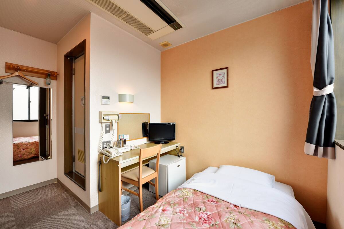 A hotel room with a bed, a desk with a computer, a small fridge, a mirror, and a window with curtains.