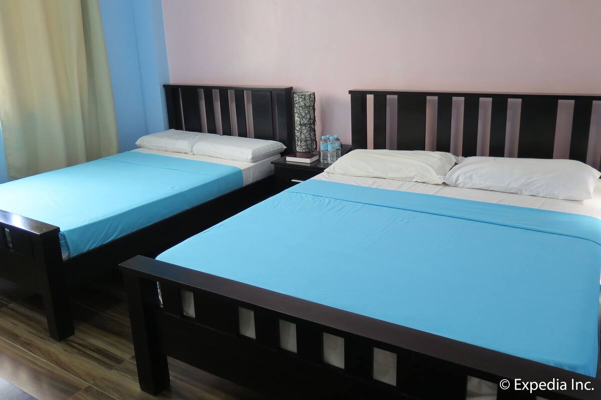 Dos camas individuales con estructura de madera y ropa de cama azul, una mesita de noche con una botella de agua y un jarrón decorativo.