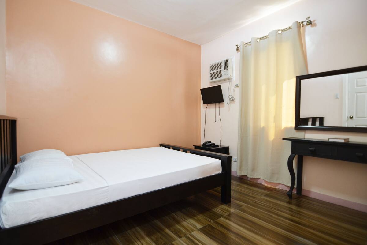 Una cama individual con estructura de madera, una mesita de noche, un televisor instalado en la pared y un espejo sobre un tocador.