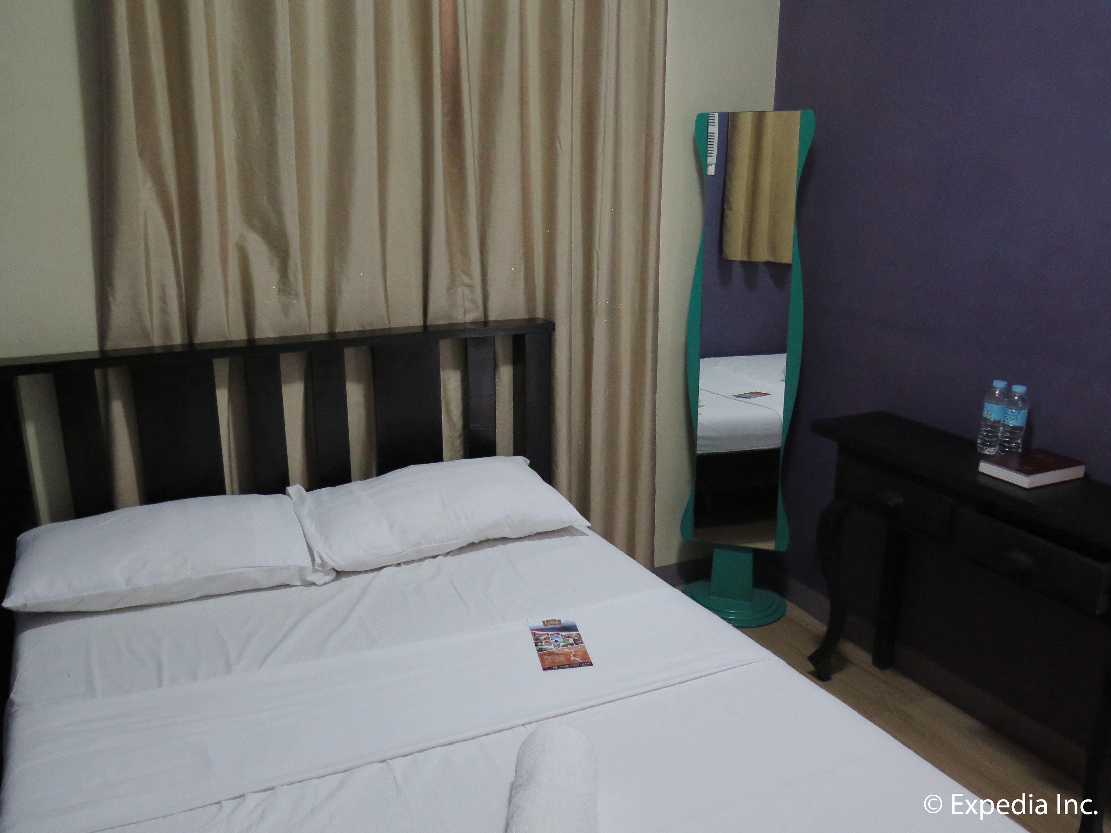 A hotel room with a bed, a nightstand with a book and water bottles, a mirror, and a window with curtains.