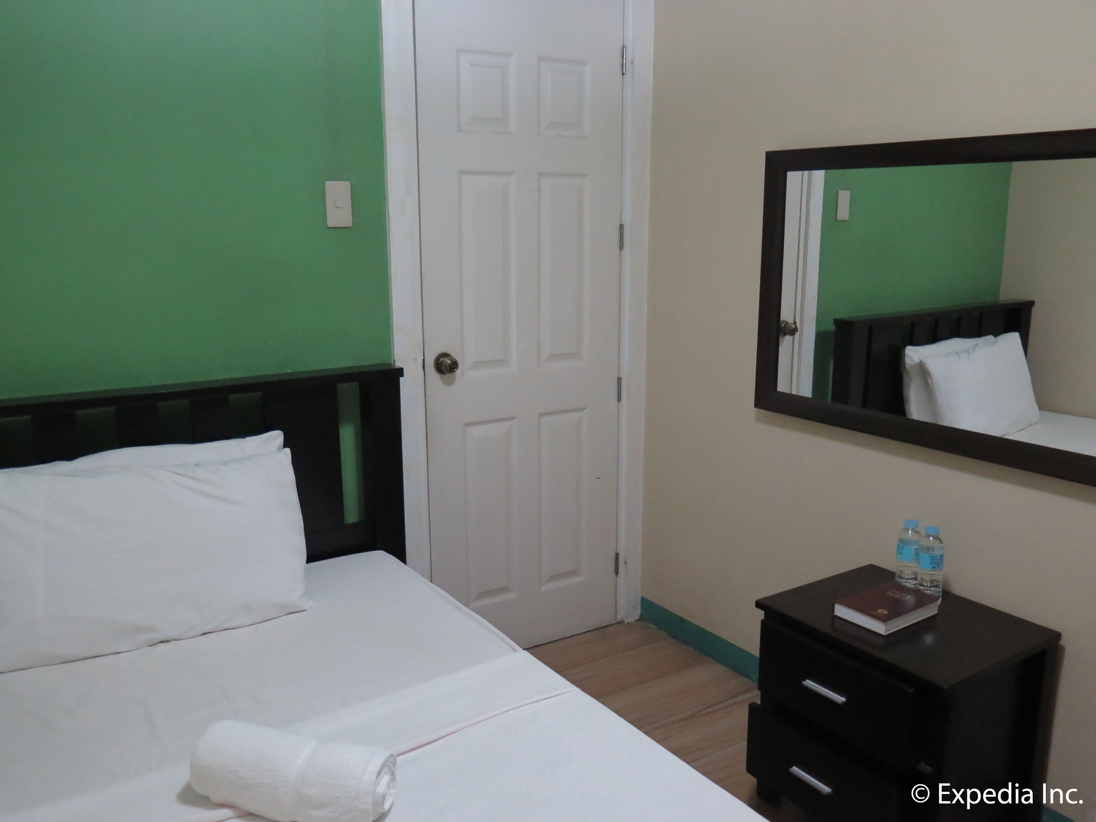 A hotel room with a bed, a bedside table with a book and water bottles, a mirror, and a door.