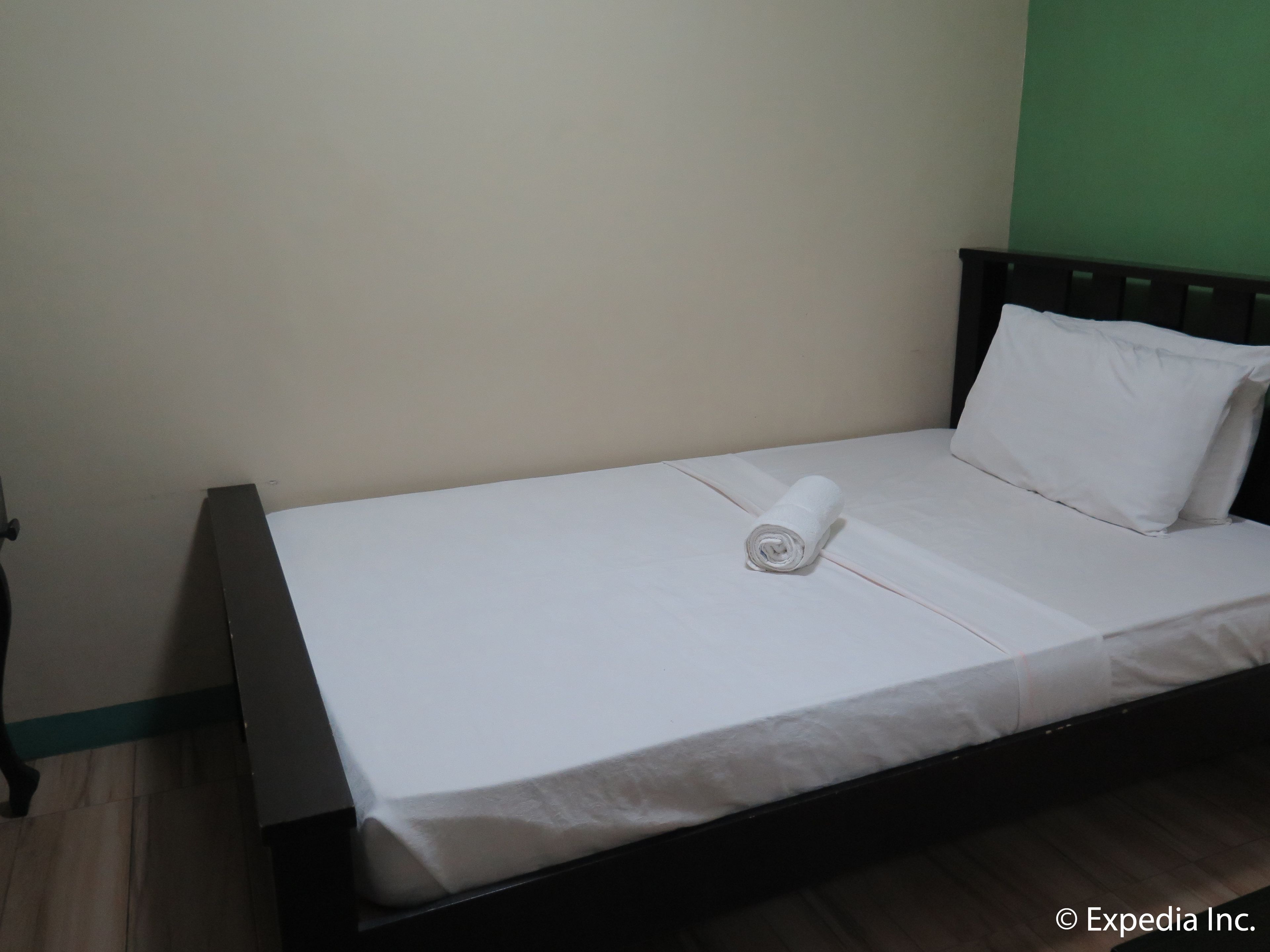 A single bed with a white mattress and pillow, a wooden headboard, and a green wall in the background.